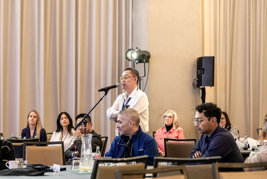 An attendee speaks into a microphone during a conference discussion while other participants listen from seated tables.